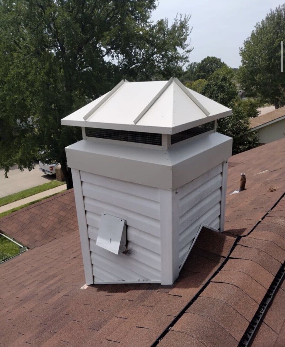 White metal chimney cap with hipped top and mesh screen on a white sided chimney chase above a brown shingle roof