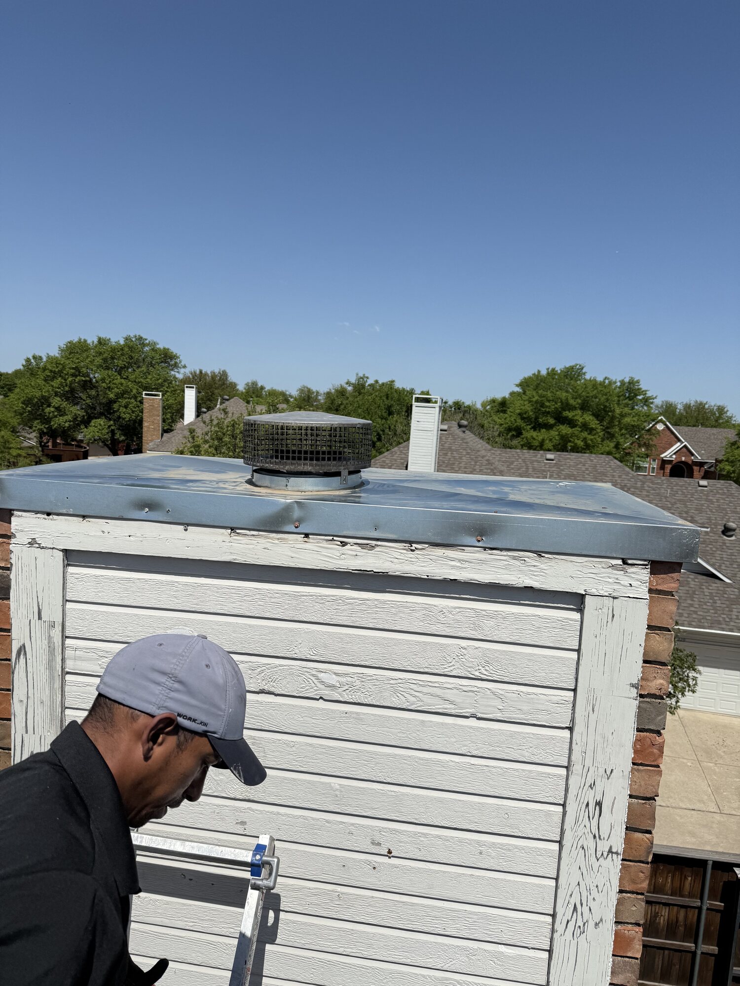 Technician working at the top of a chimney chase beside a metal chase cover with a round flue rain cap