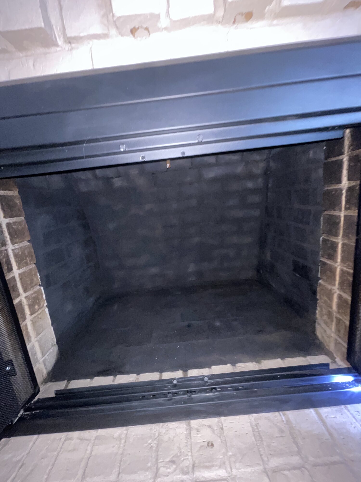 Interior of a masonry fireplace firebox with soot-darkened brick walls behind an open metal fireplace door frame