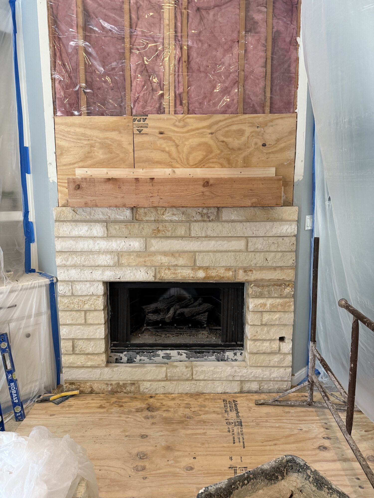 Fireplace at an early renovation stage with the upper wall opened, exposing wood framing, insulation and plywood above the stone surround