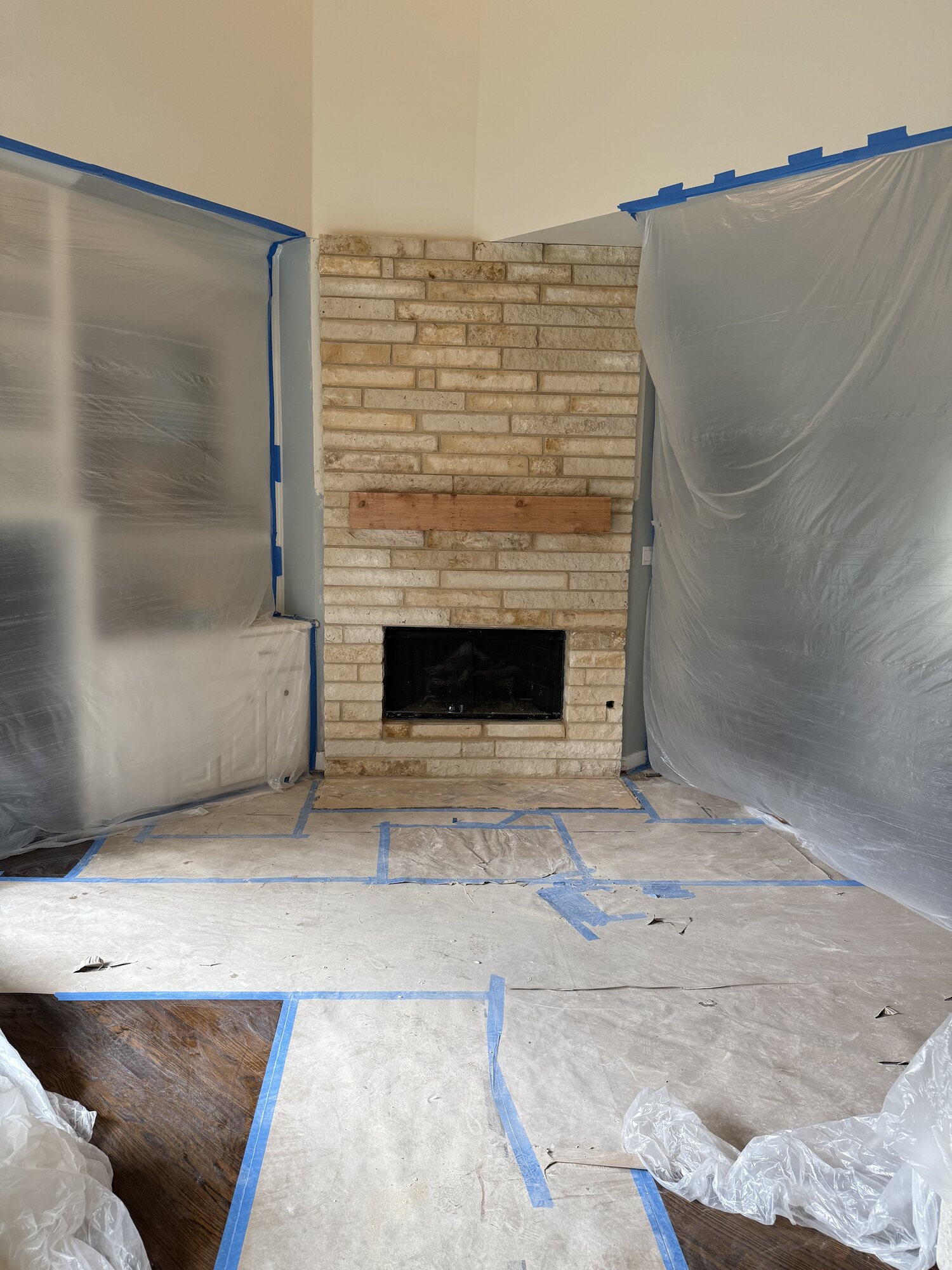 Fireplace renovation in progress with the room masked in plastic sheeting and a bare wood mantel board set on the stone face
